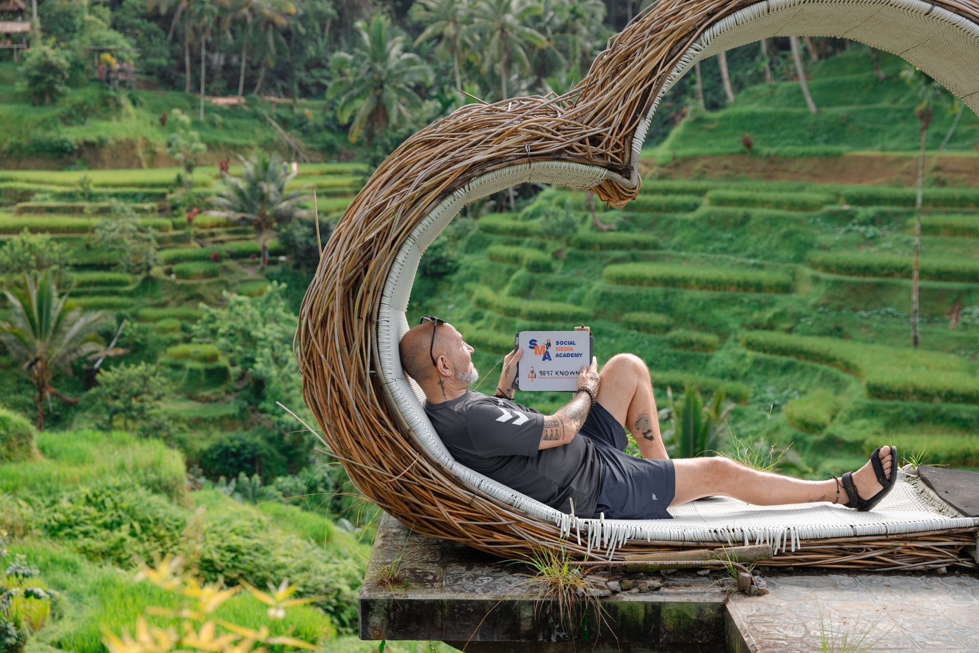 Man relaxing with a tablet in a heart-shaped wicker seat overlooking green rice terraces.