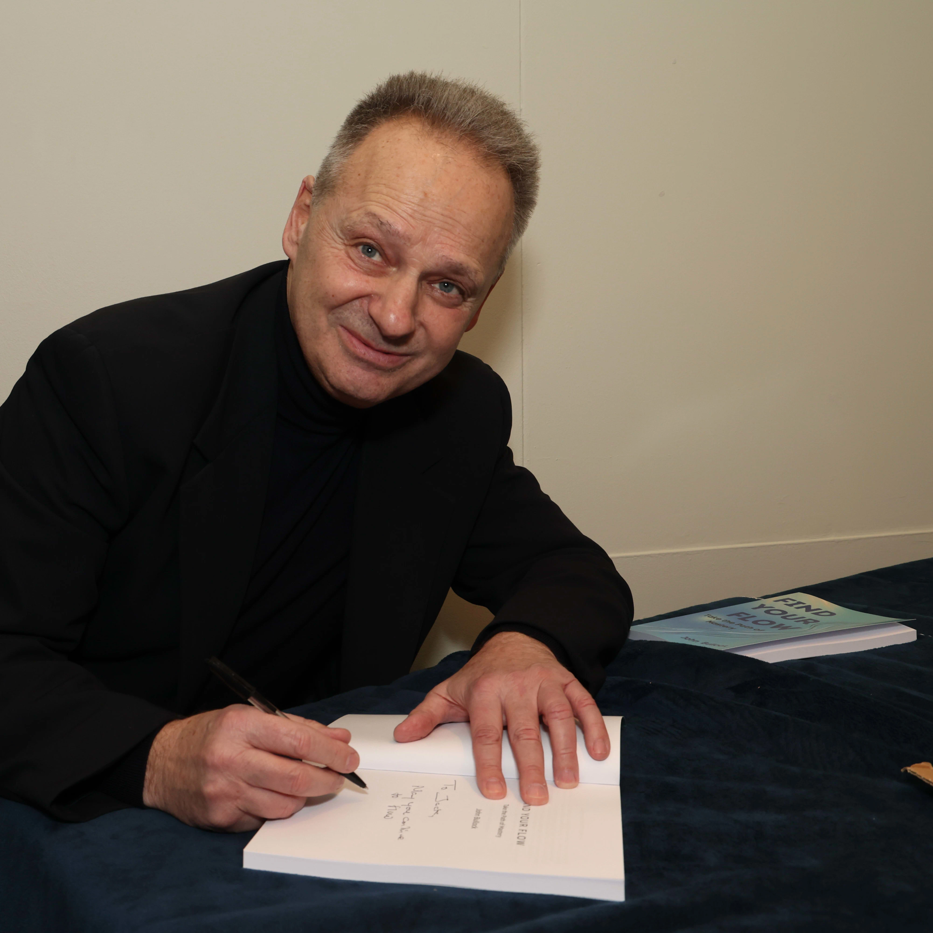 Man in black jacket signing a book at a table; neutral expression.