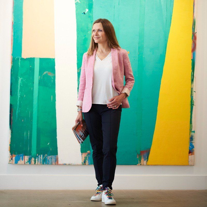 Woman in pink blazer stands in front of colorful abstract art, holding a magazine.