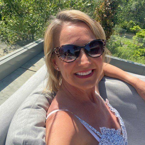 Woman in sunglasses smiles, sitting on outdoor furniture. Sunny day, greenery in background.