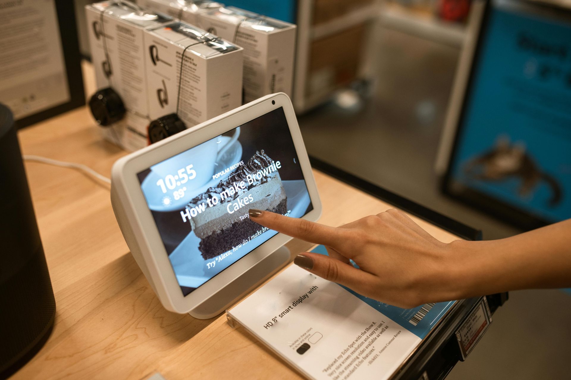 A person's hand touches an Echo Show screen displaying a cake recipe in a store setting.