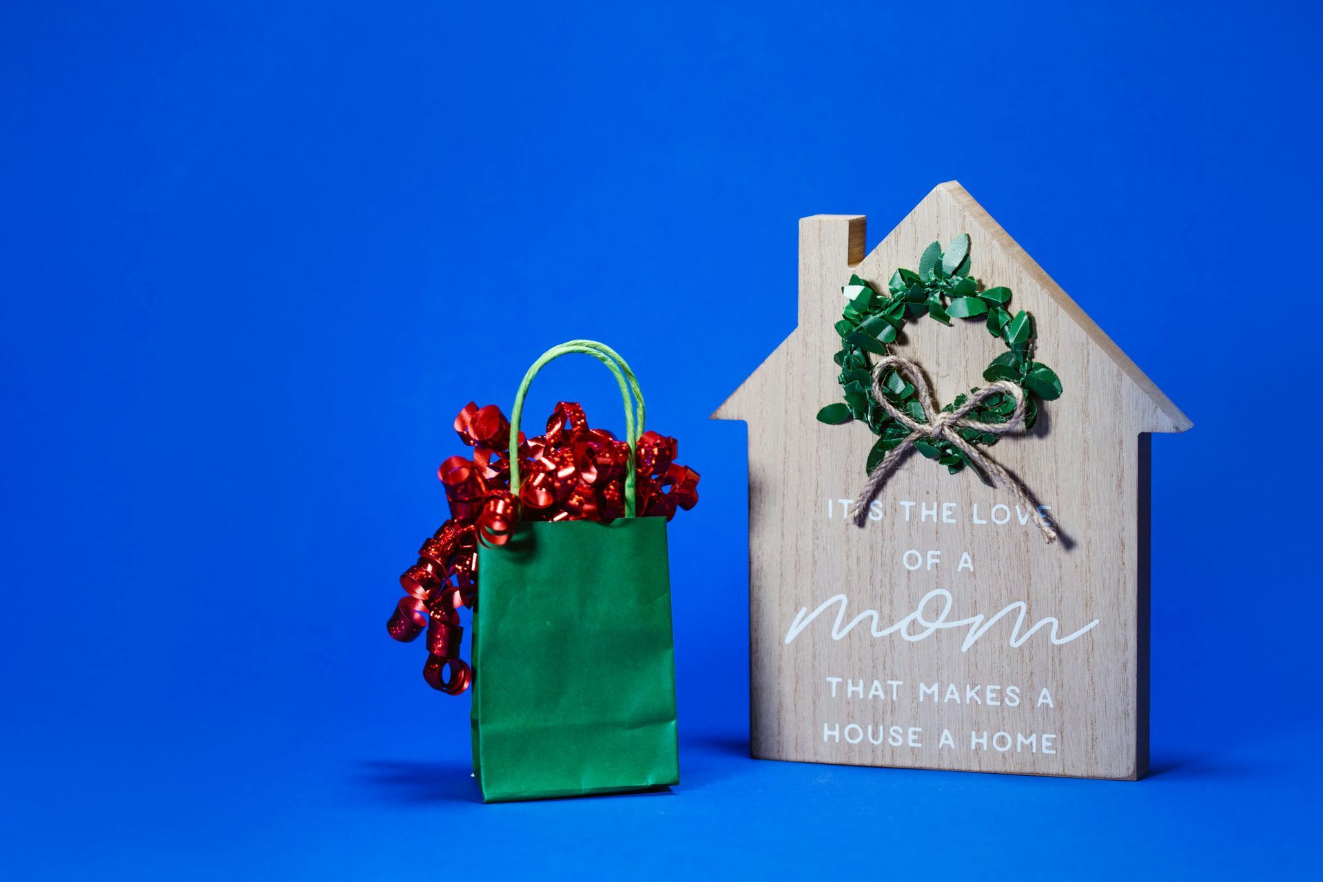 Green gift bag with red bow, and wooden house decoration with a wreath, against a blue backdrop.