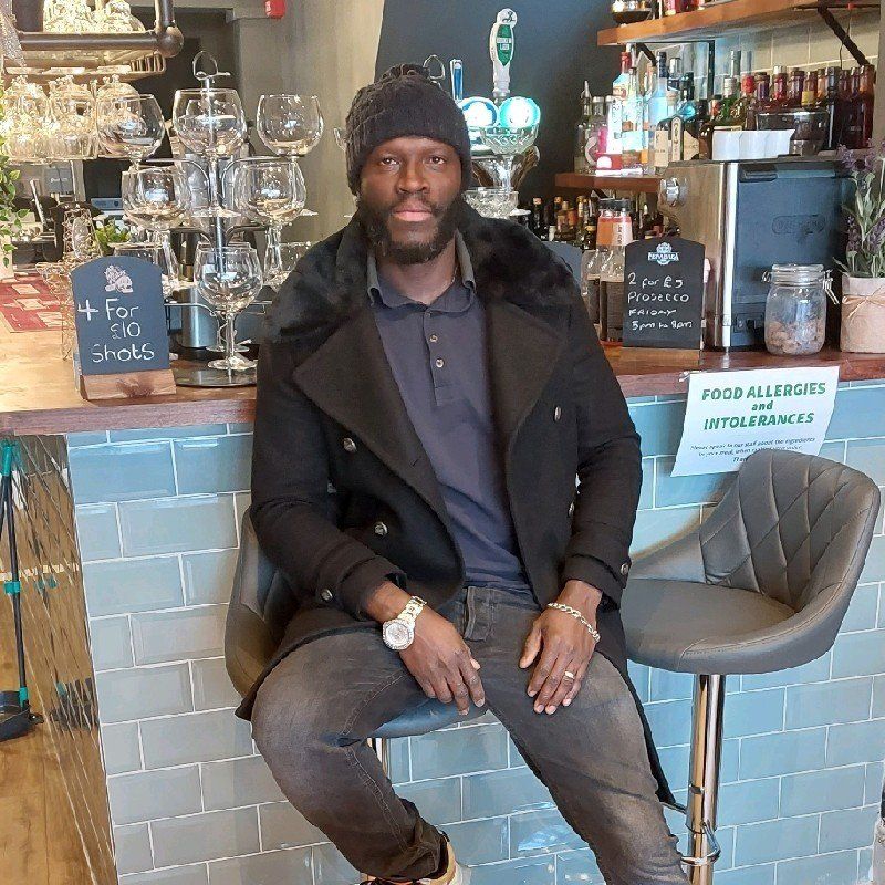 Man seated at a bar, wearing black coat and hat, looking at the camera. Bar setting.