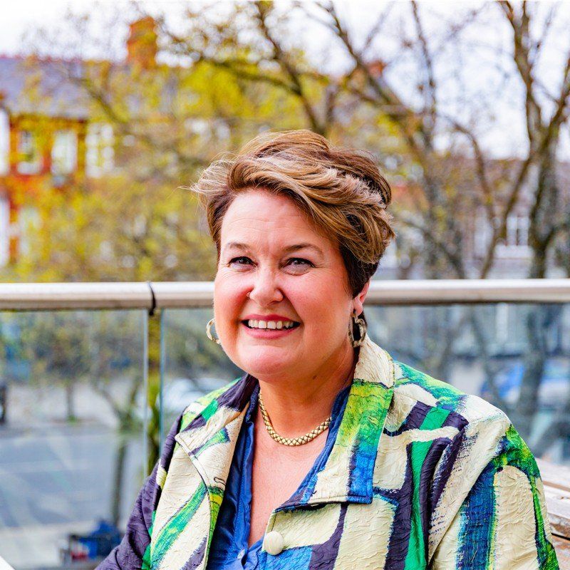 Woman in patterned jacket smiles, outdoors with building background.