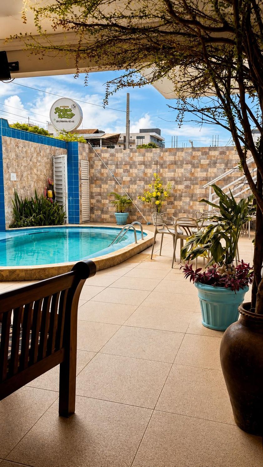 Pátio com uma pequena piscina azul, muro de pedra, vasos de plantas e cadeiras de exterior sob um telhado sombreado.