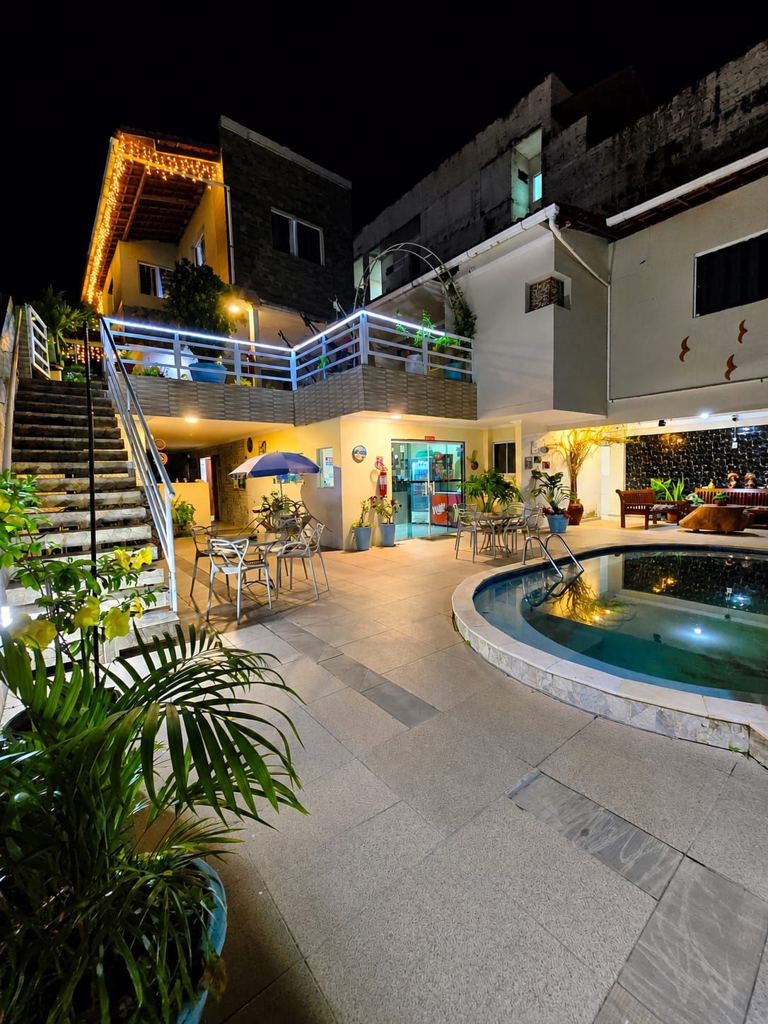 Pátio noturno com piscina, escadas iluminadas e um edifício de dois andares com varandas.