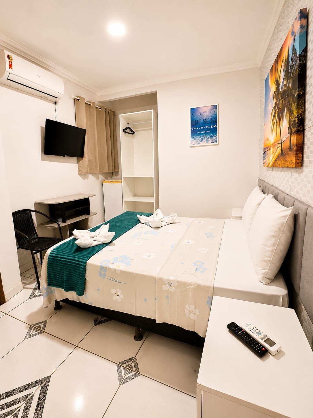 Quarto pequeno com cama, guarda-roupa aberto, cadeira de escritório, prateleira de parede e piso de azulejo.