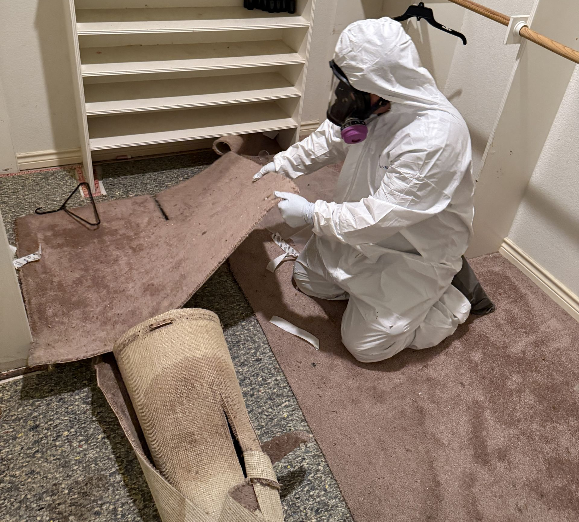 Person in white protective suit kneeling on carpet, examining a damaged floor panel in a hallway.