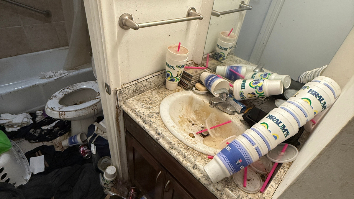 Cluttered bathroom sink and counter with spilled tubes and toiletries, cabinet drawer open, toilet visible in background