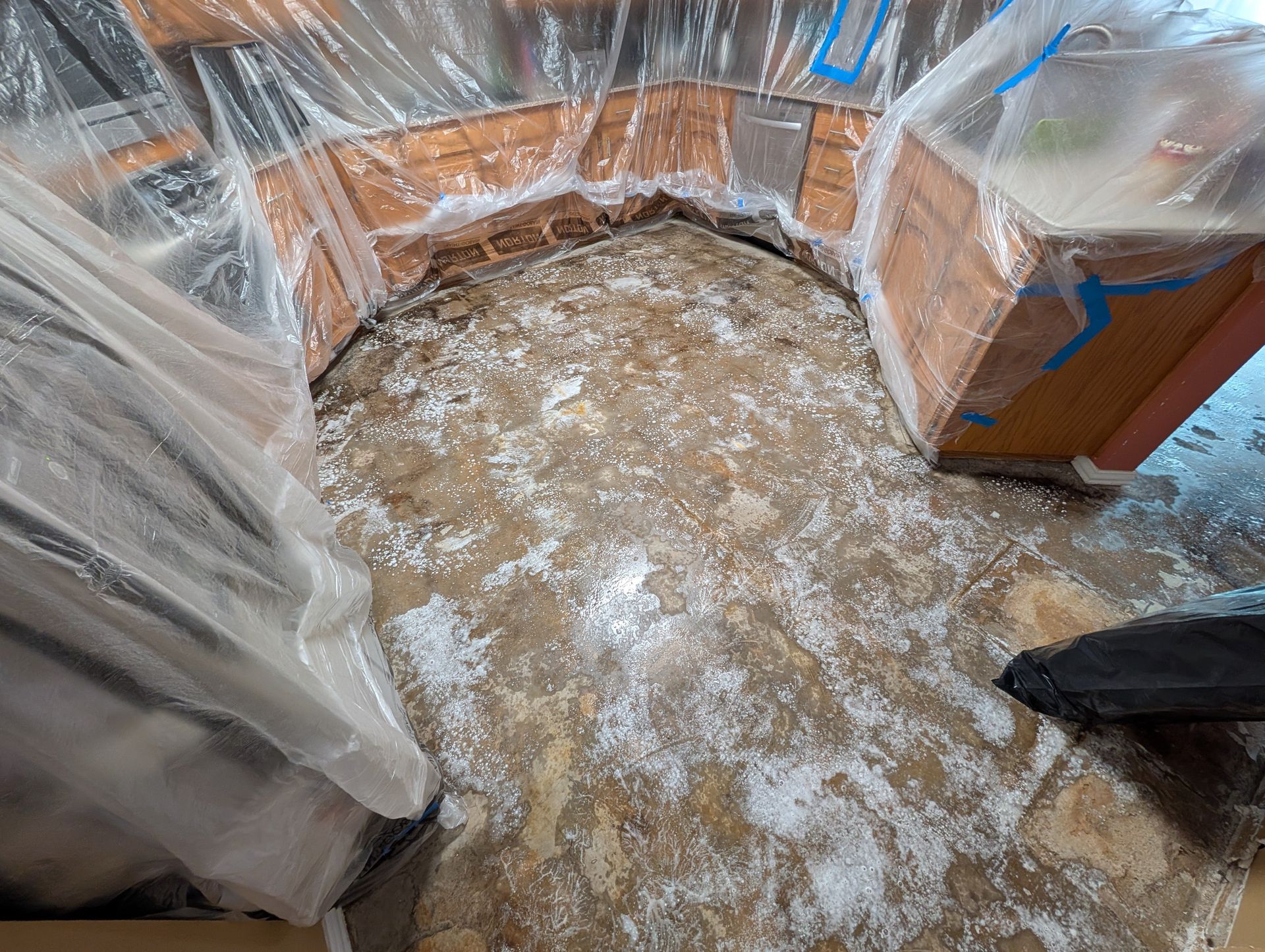 Kitchen floor under renovation, with cabinets covered in plastic sheets and concrete flooring exposed and dusty.