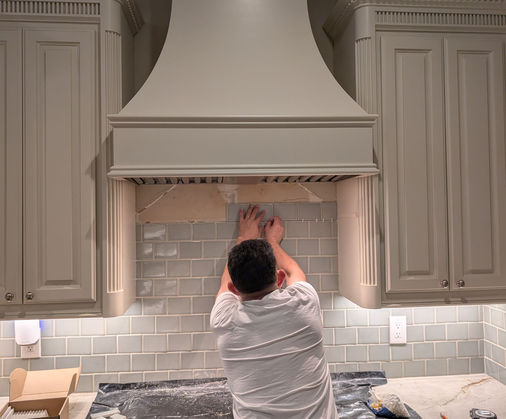 Person adjusting tile backsplash beneath a white range hood in a kitchen