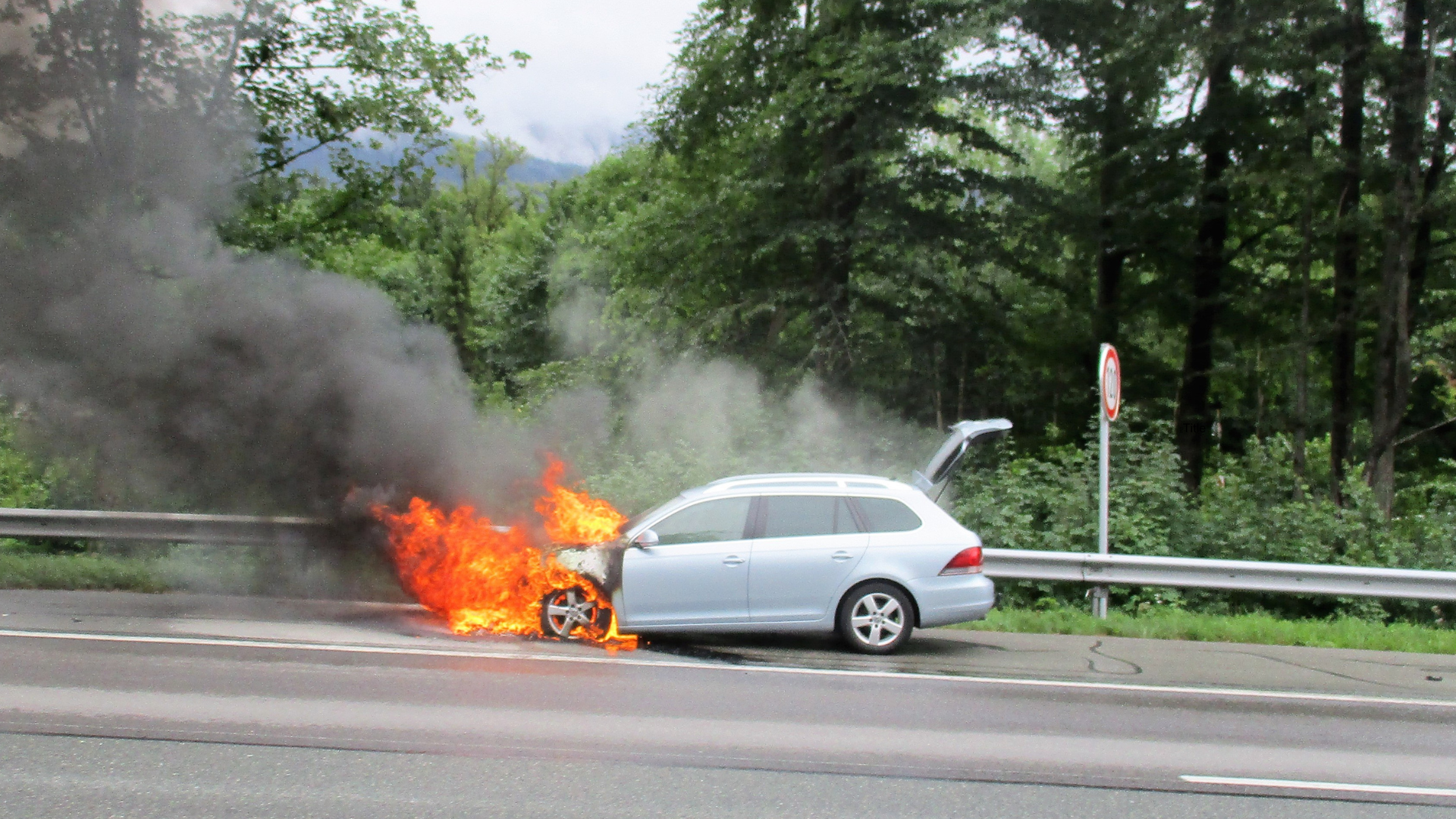 Car engulfed in flames on roadside, emitting black smoke. Trunk open.