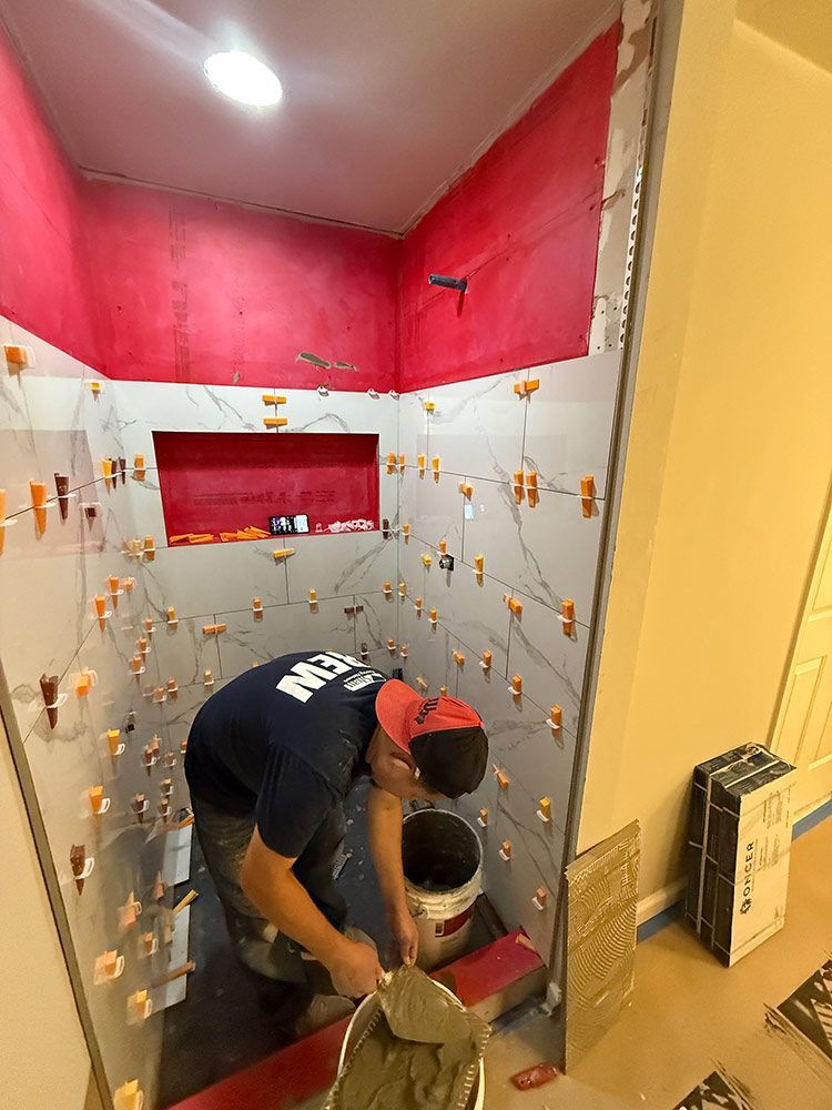 Person tiling a shower stall with white tiles, using orange tile spacers. Red walls are visible.