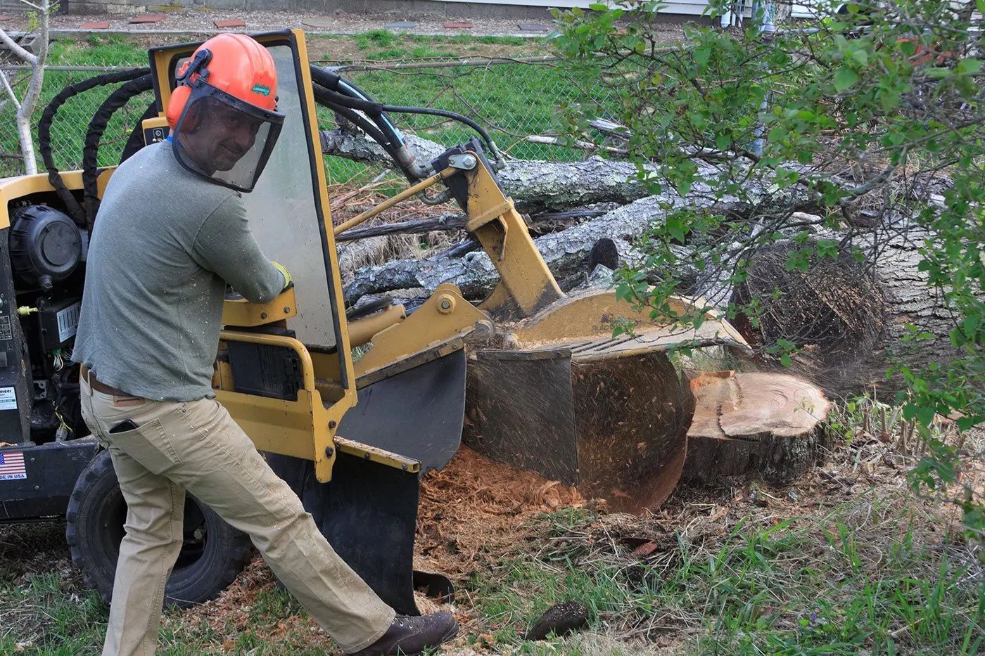 Man operating a yellow stump grinder on a tree stump, with safety gear in a yard.