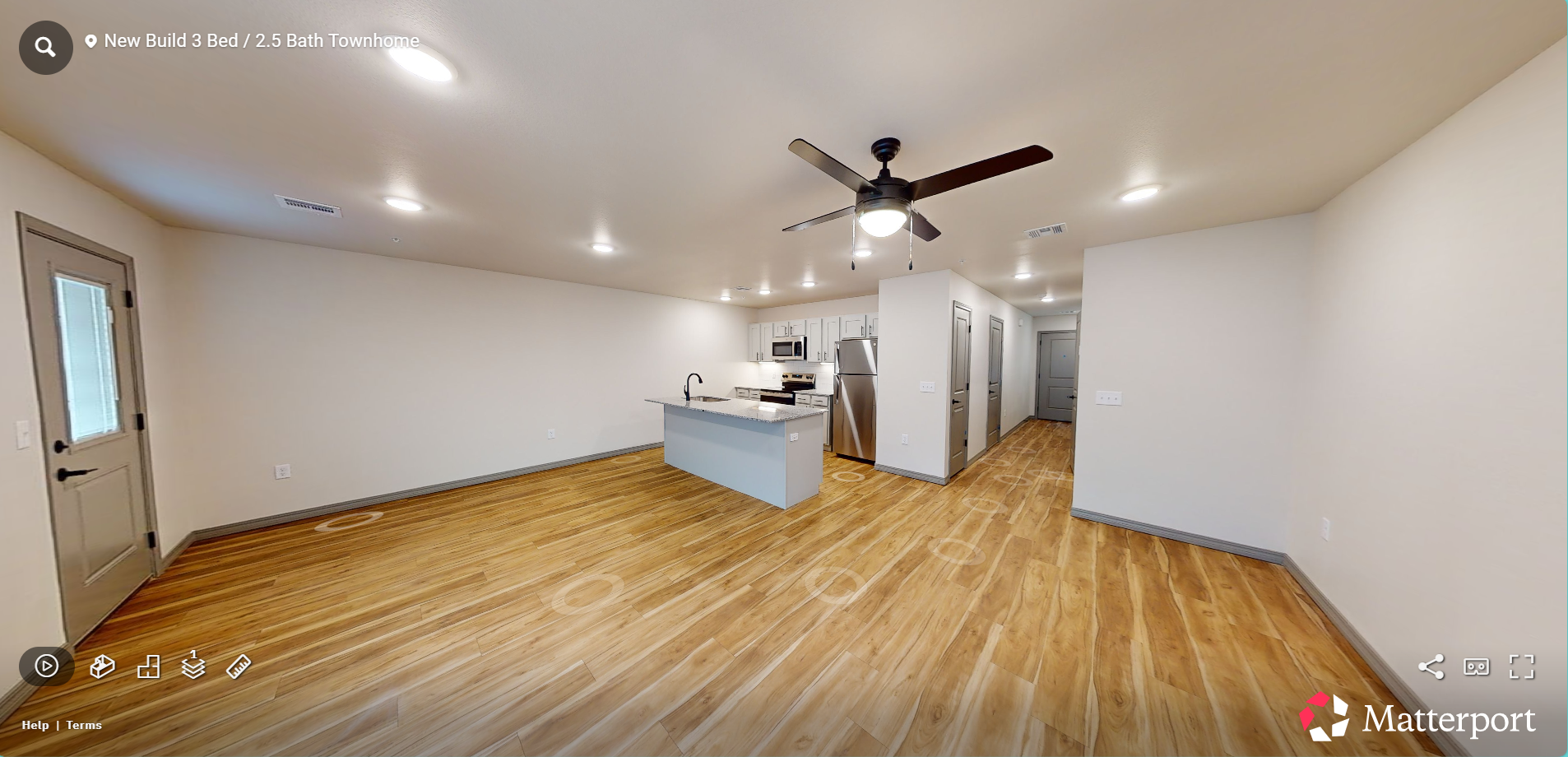 Empty modern living room with light wood floors, white walls, ceiling fan, and open kitchen island