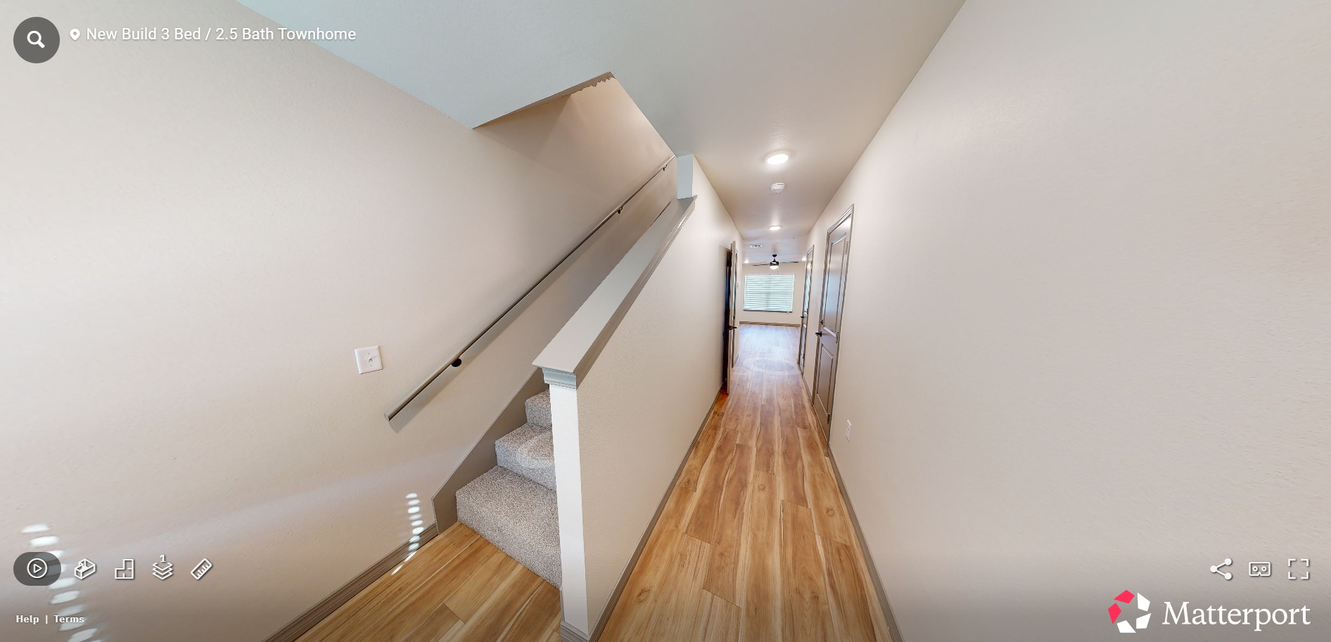 Narrow hallway with wood floors, white walls, staircase railing, and bright light at the far end