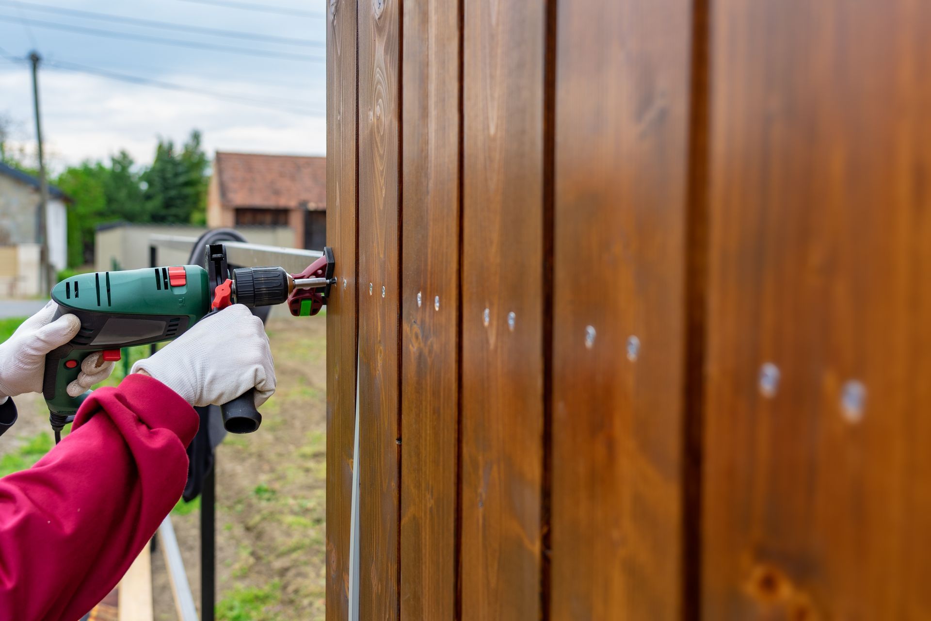 Person wearing gloves using a drill to install something on a stained wooden fence
