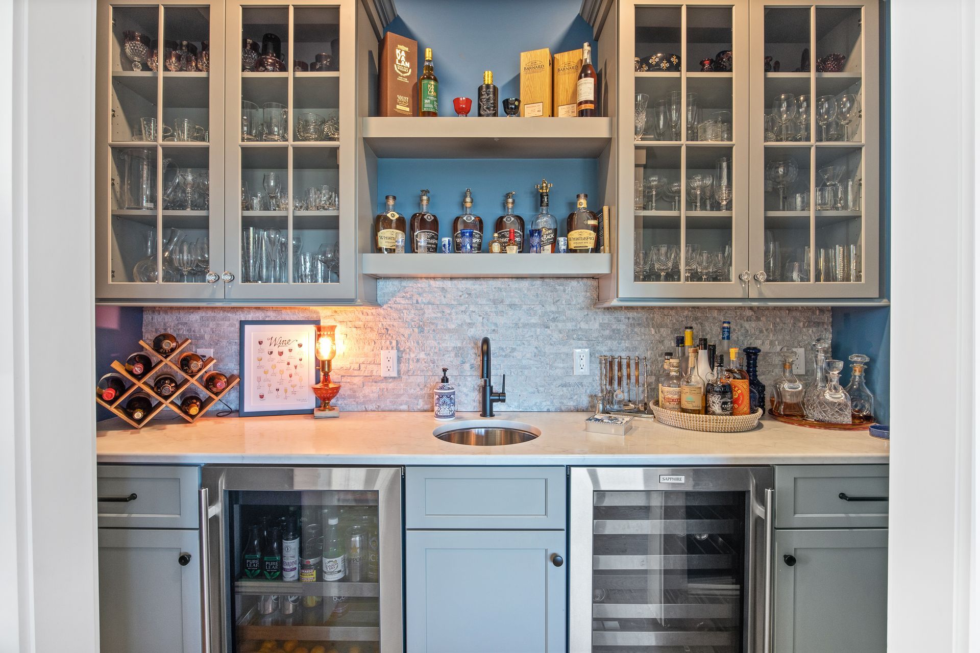 Built-in home wet bar with glass cabinets, blue-gray cabinetry, white marble countertop, and bottles displayed inline