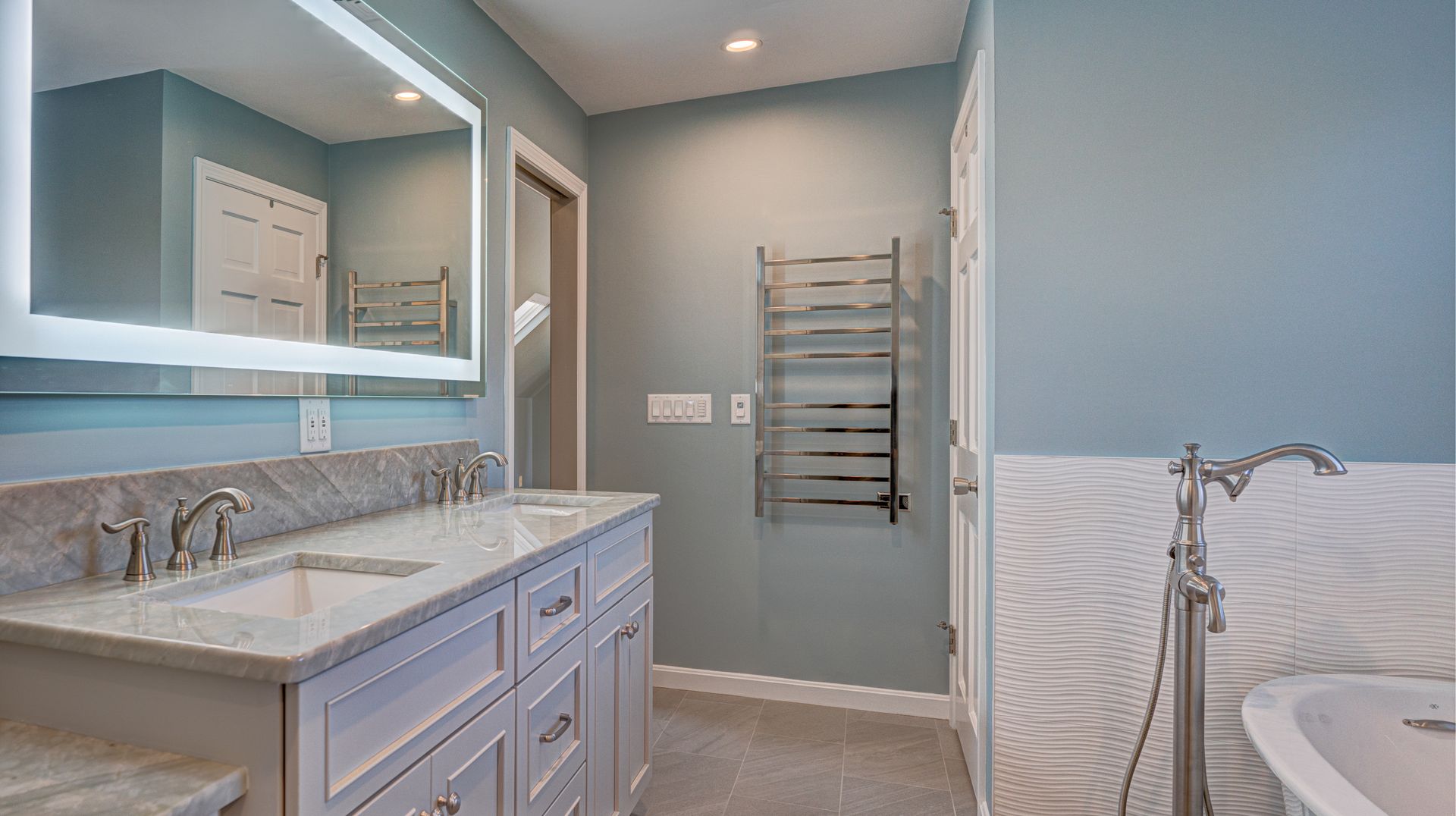 Modern bathroom with double vanity, large mirror, towel rack, and white tub with blue walls.