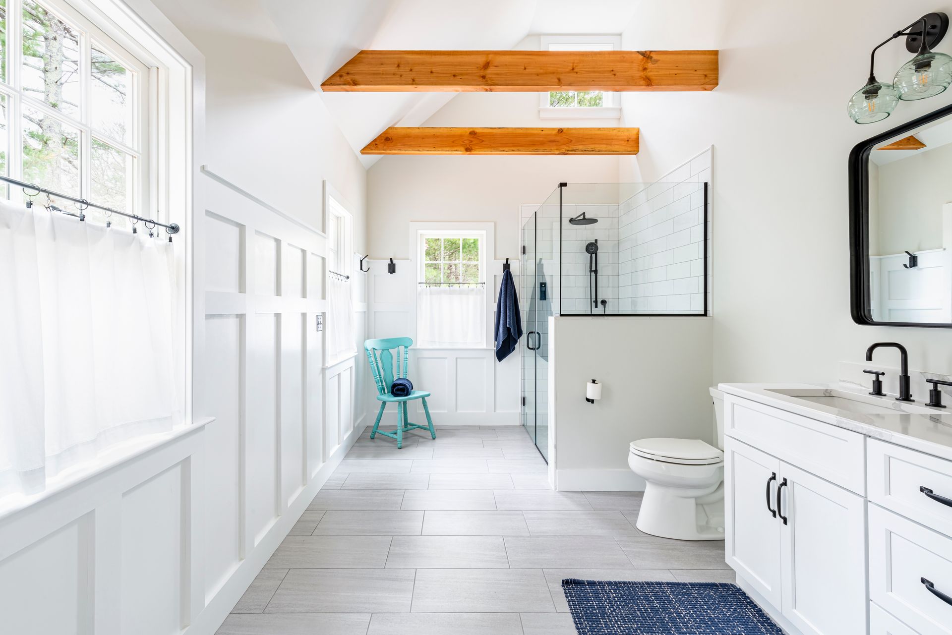 Bright white bathroom with wood ceiling beams, double vanity, freestanding tub, and teal chair near the window