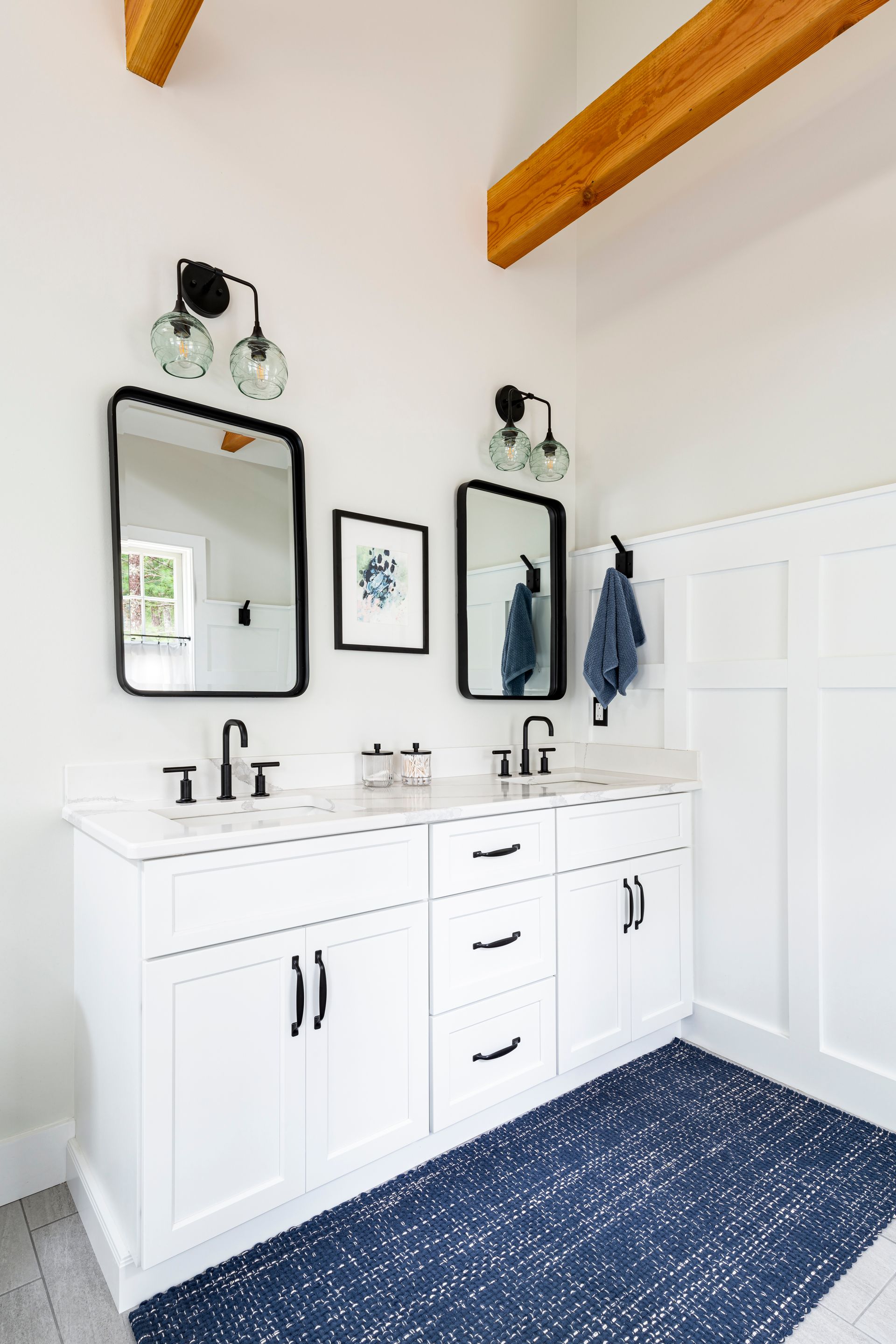 Bright white bathroom vanity with double sinks, black mirrors, navy rug, and wood ceiling beams