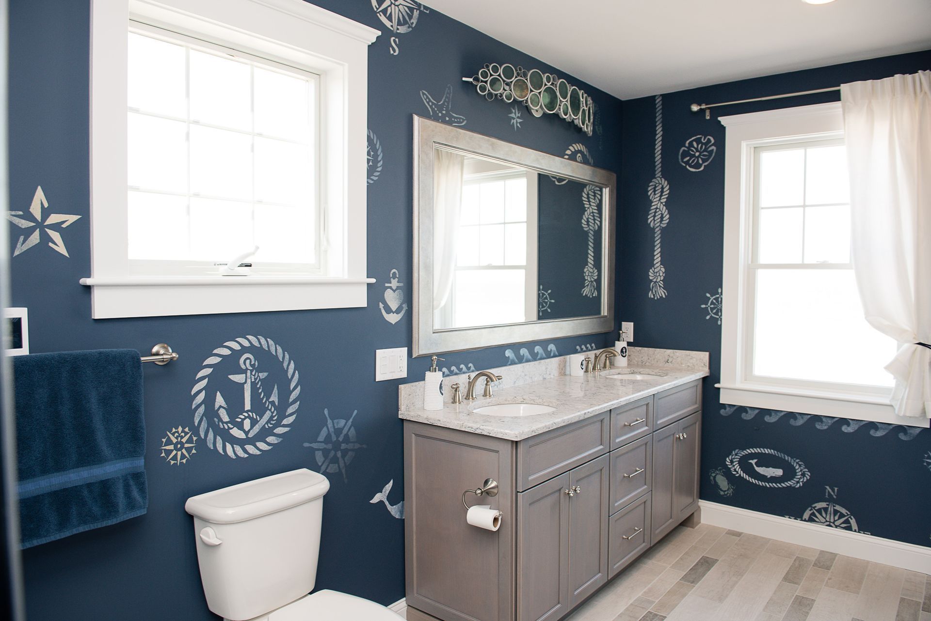 Blue bathroom with white nautical wallpaper, double vanity, mirror, windows, and toilet