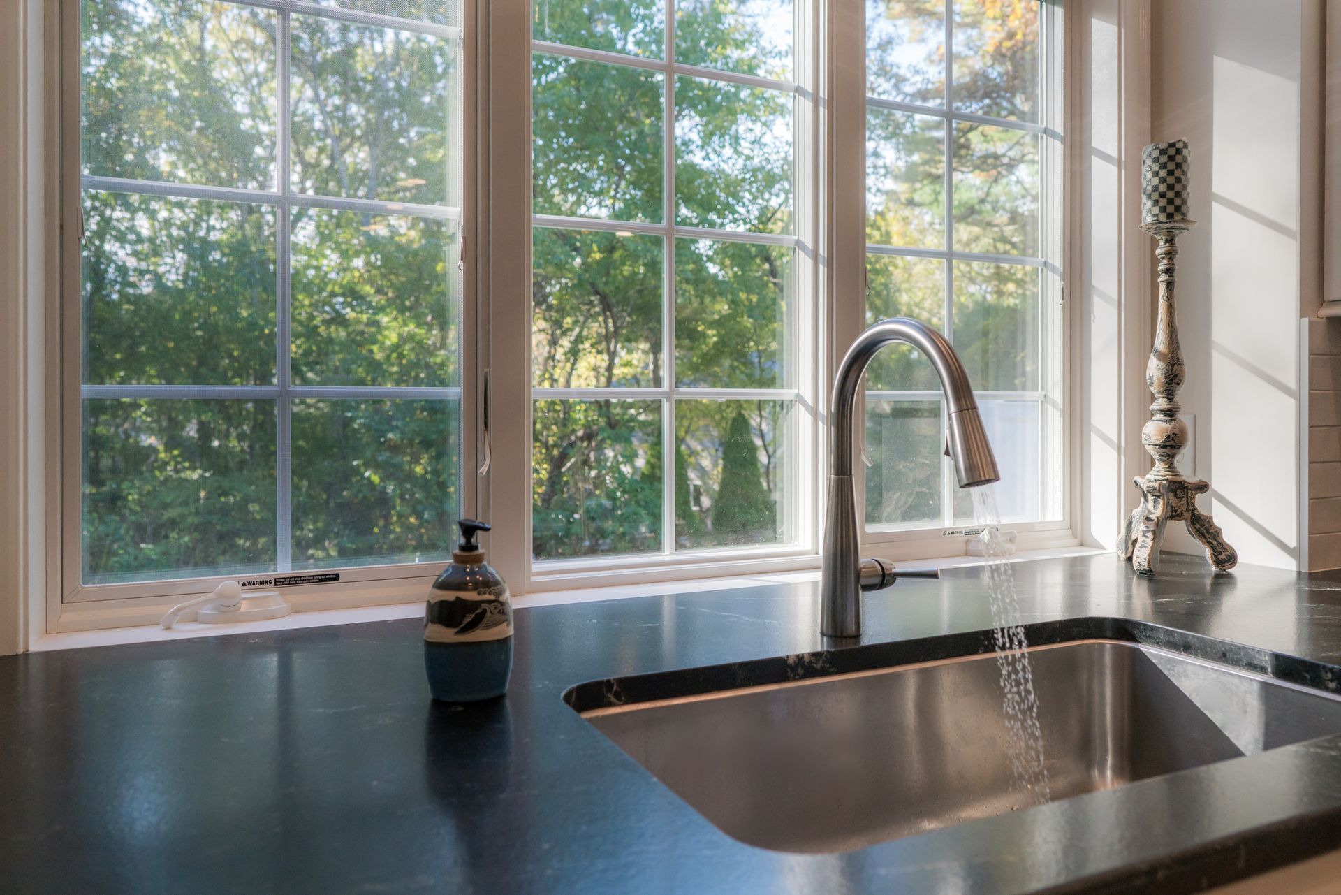 Bright kitchen sink by a large window overlooking green trees, with dark countertop and chrome faucet