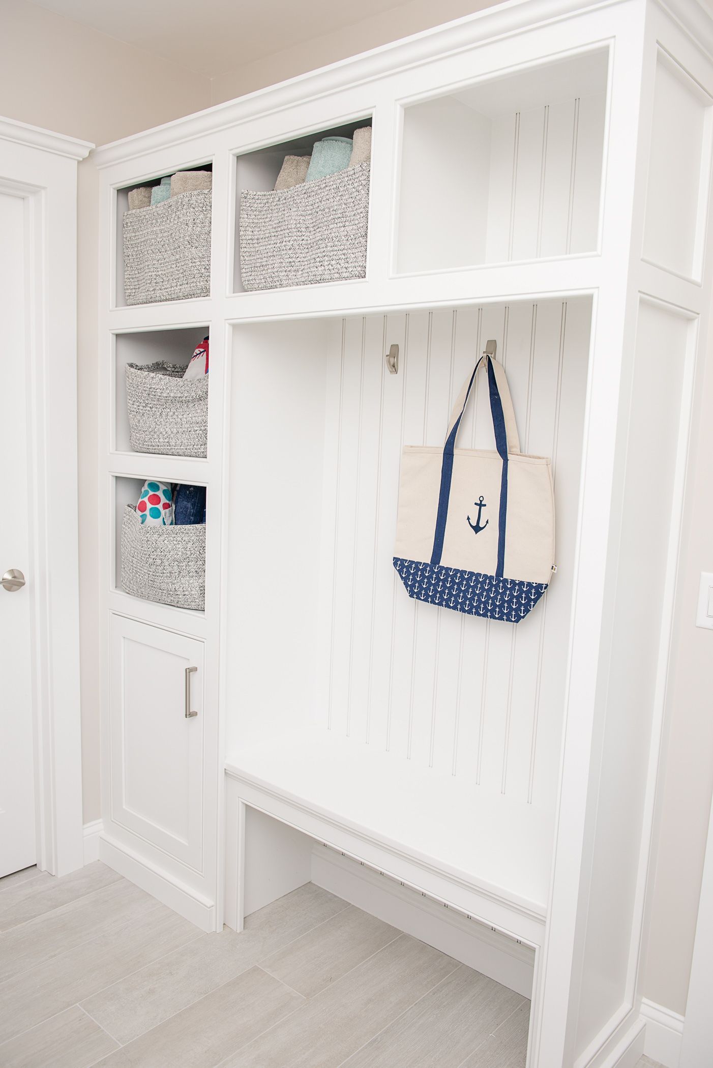 White built-in entryway cubbies with baskets, hooks, a bench, and a blue tote bag hanging on the wall.
