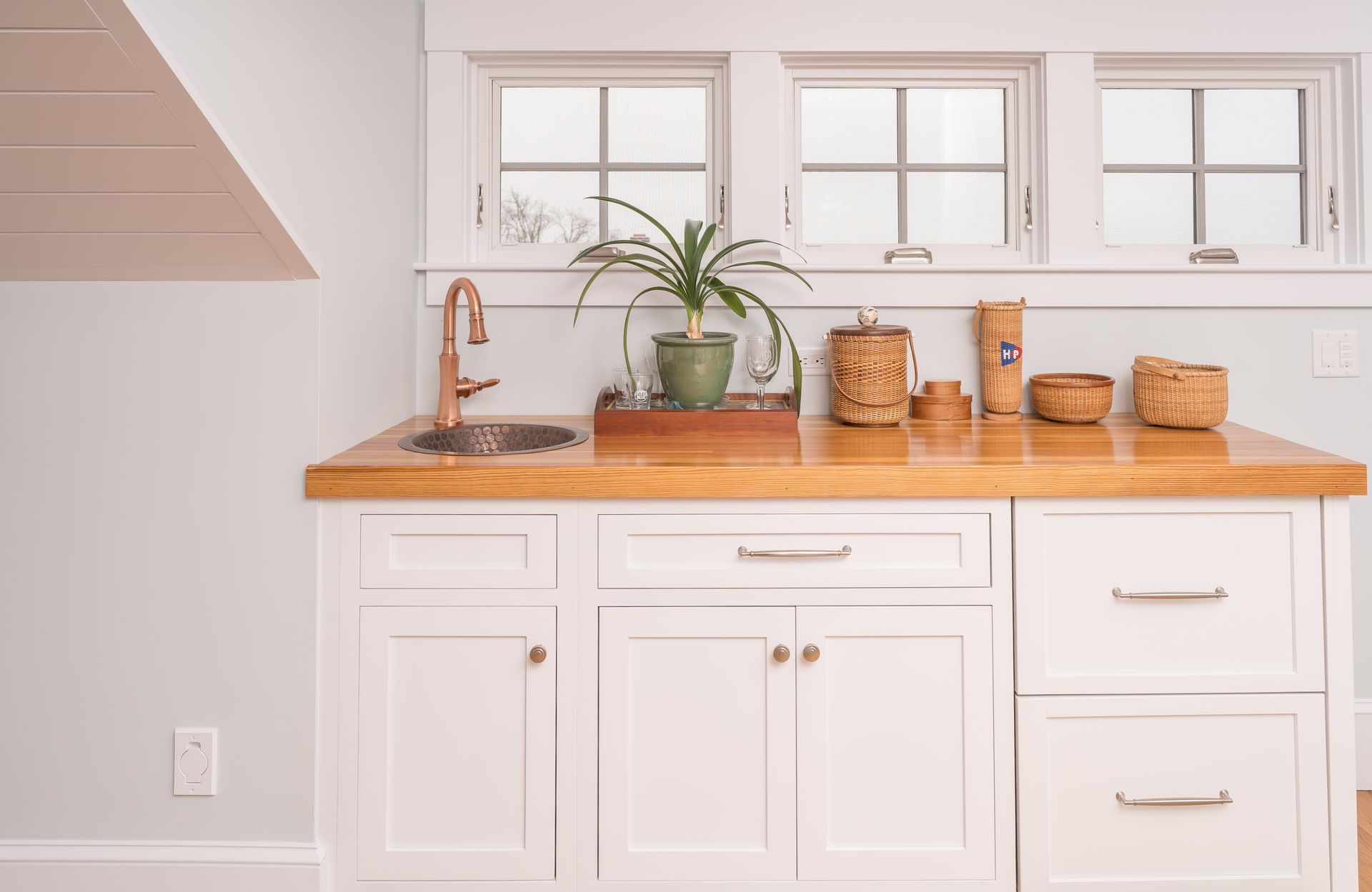 White kitchen counter with wooden top, sink, potted plant, and woven baskets beneath windowed wall cabinets