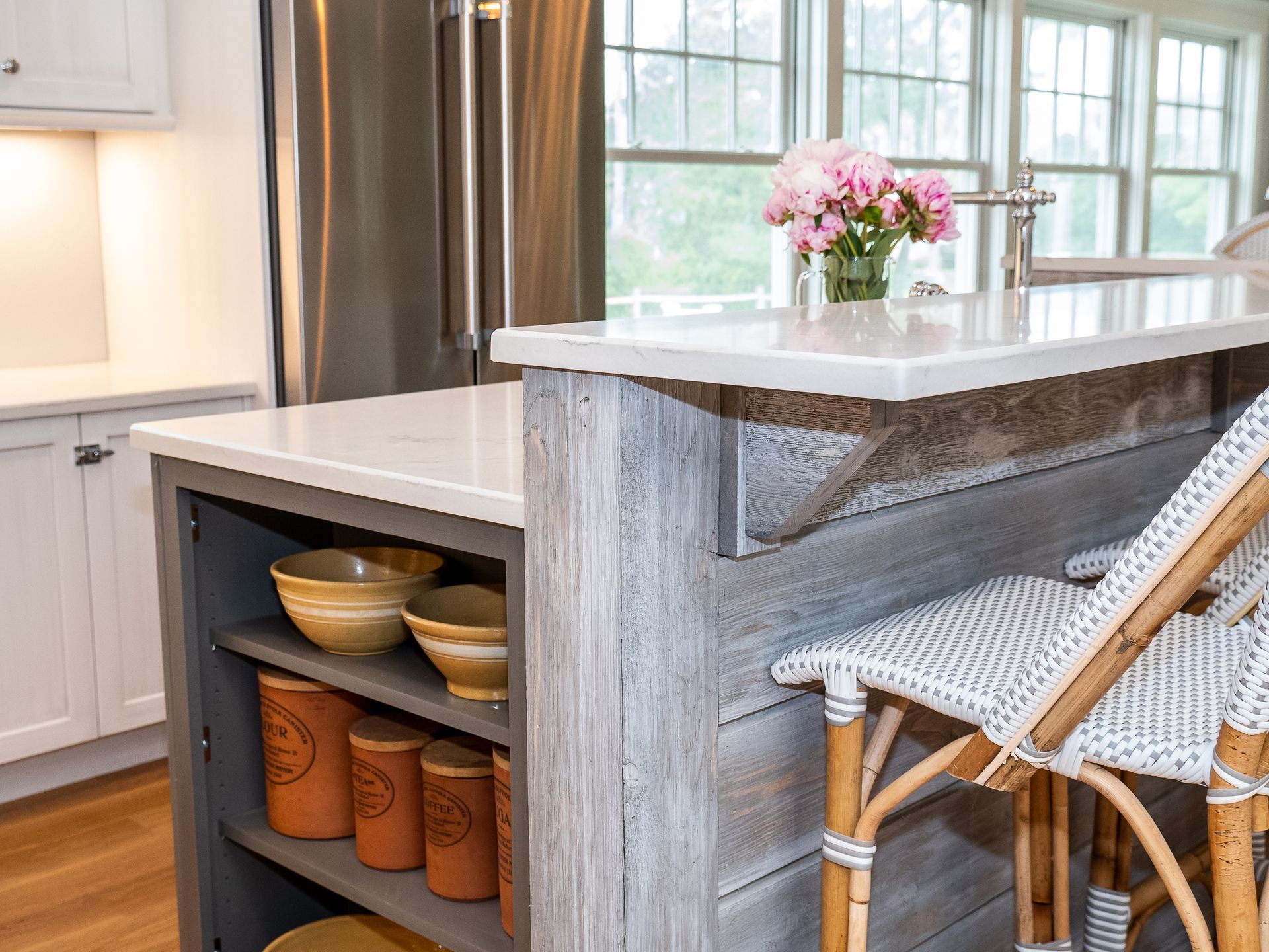 Bright kitchen island with wicker bar stools, open storage shelves, and a vase of pink flowers