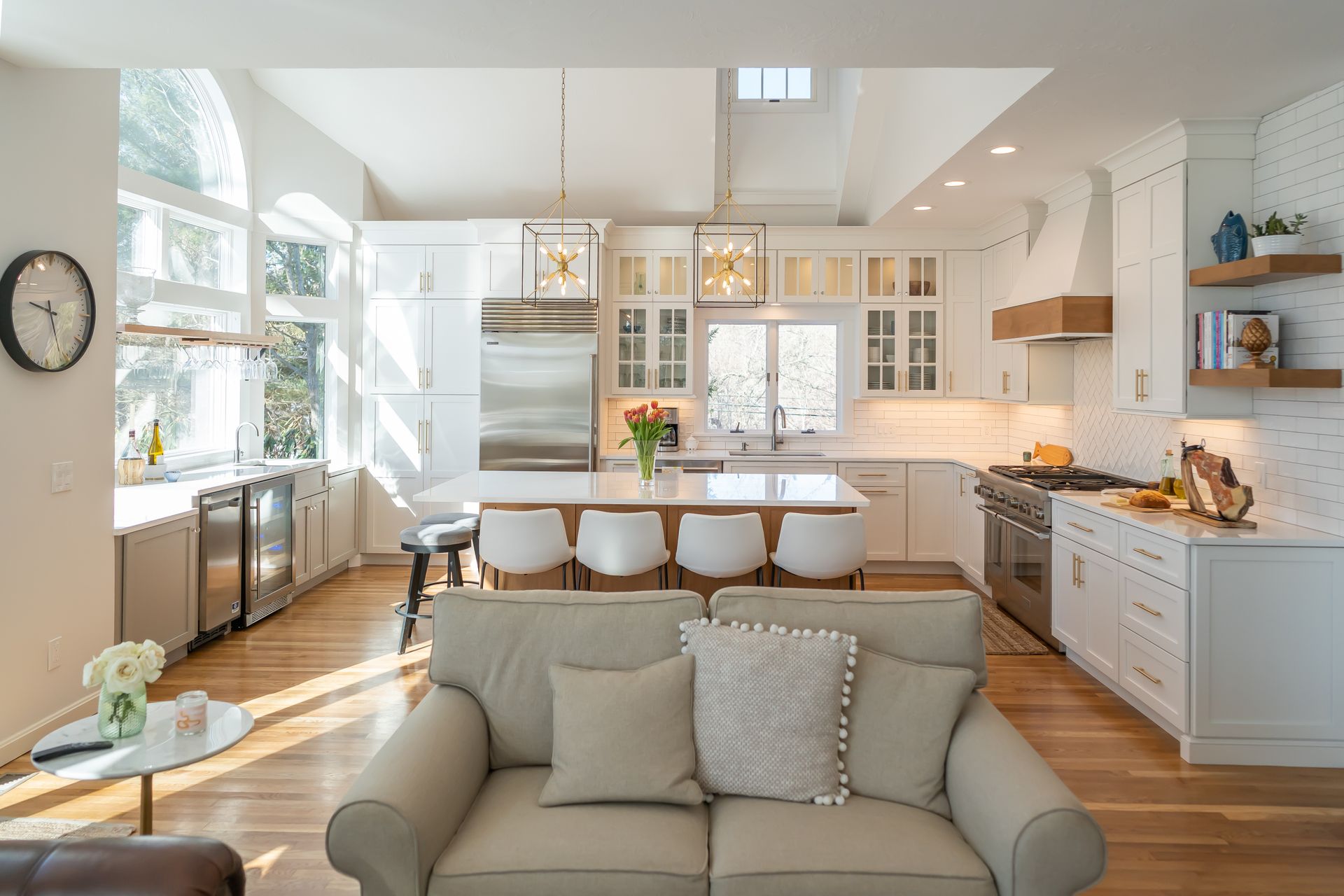 Bright modern kitchen and living area with white cabinets, island stools, and a beige sofa by large windows