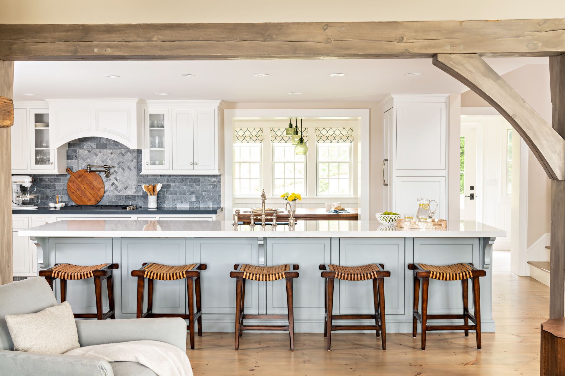 Bright farmhouse kitchen with white island, wooden stools, exposed beams, and large window lighting