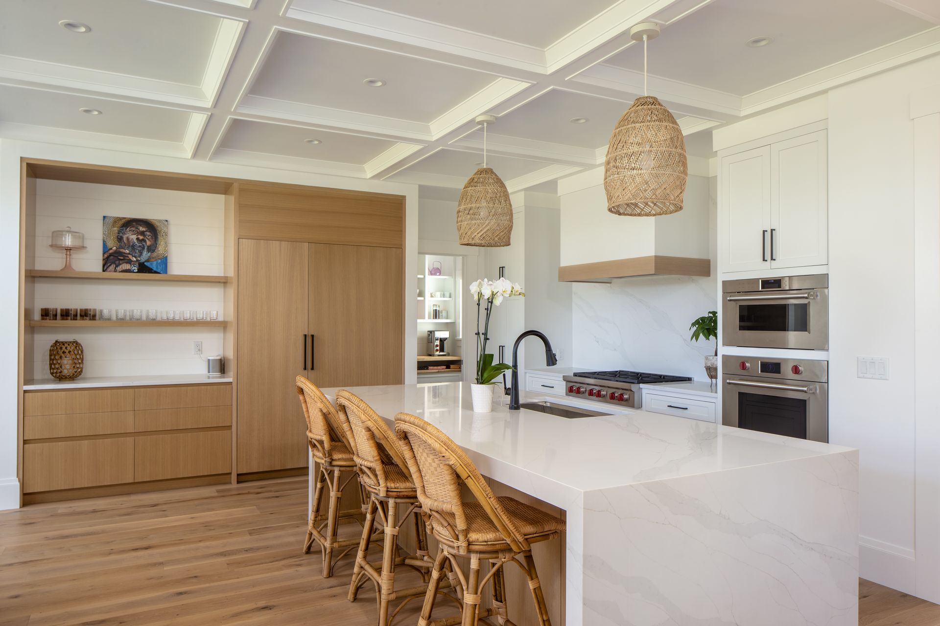 Modern white kitchen with wooden stools, island, woven pendant lights, and built-in oven and cabinets.