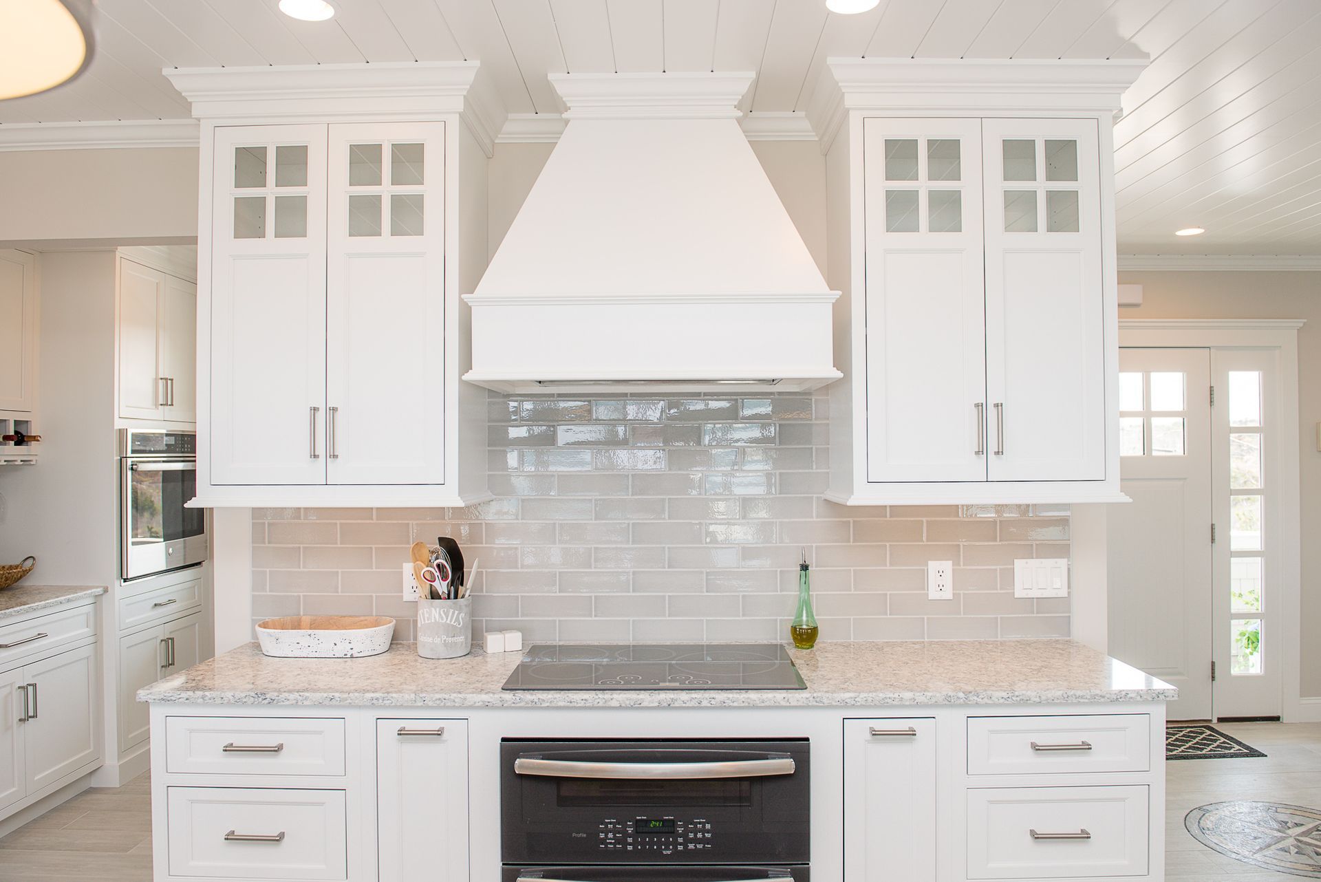 Bright white kitchen with marble countertops, glass tile backsplash, and large hood over a stove.