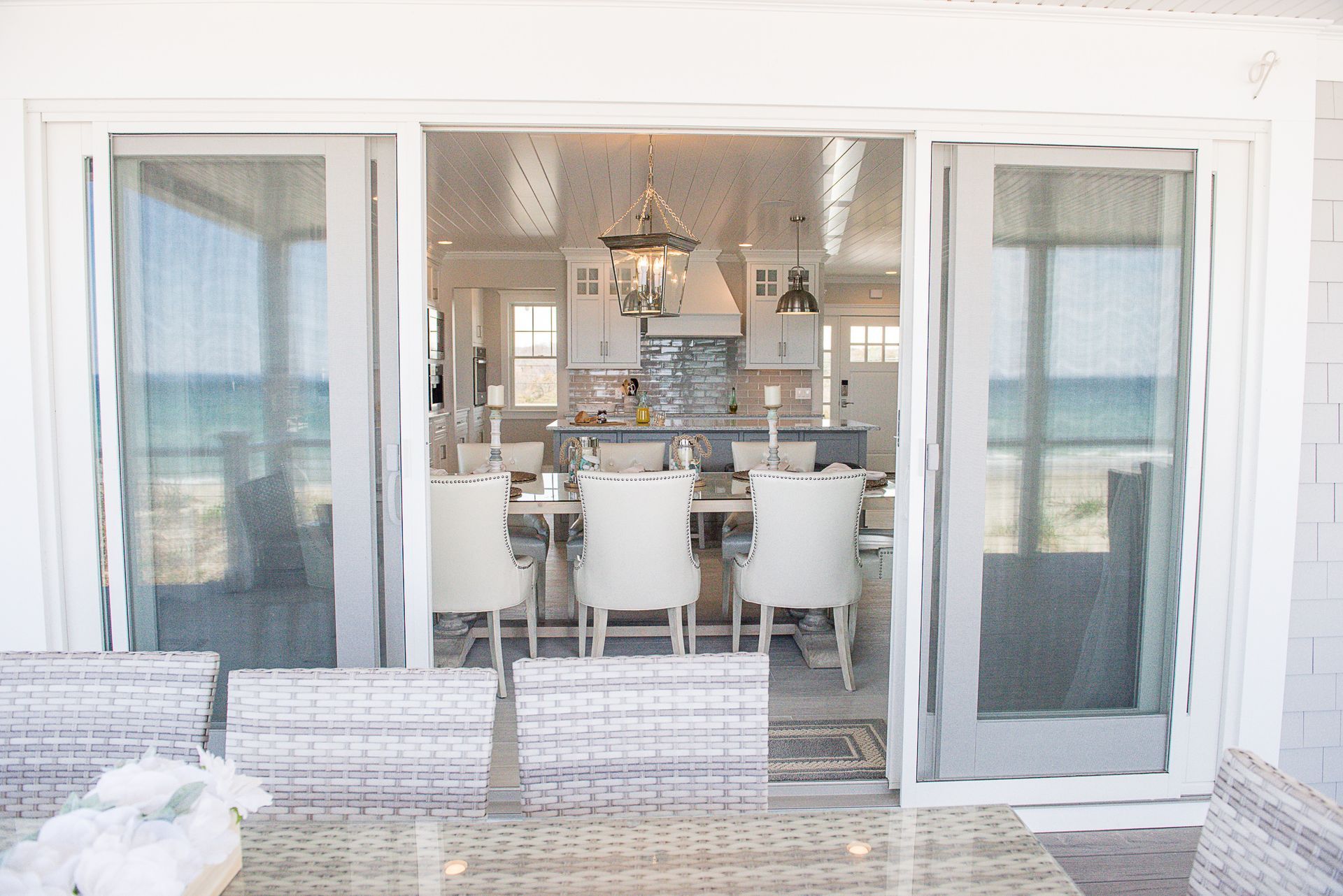 Bright seaside dining room with white chairs, chandelier, and open French doors to the ocean.
