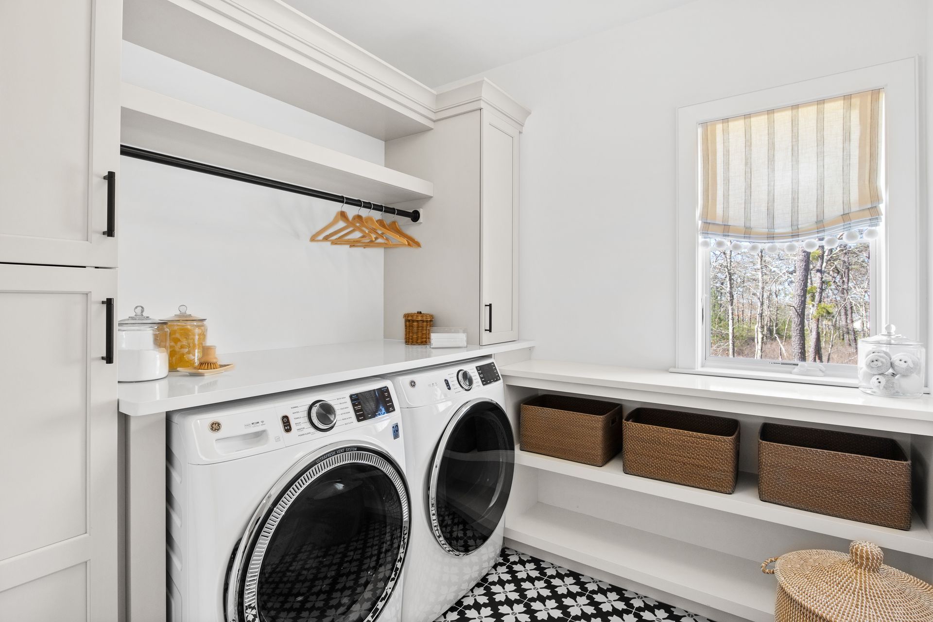 Bright laundry room with front-loading washer and dryer, white cabinets, and window bench with baskets