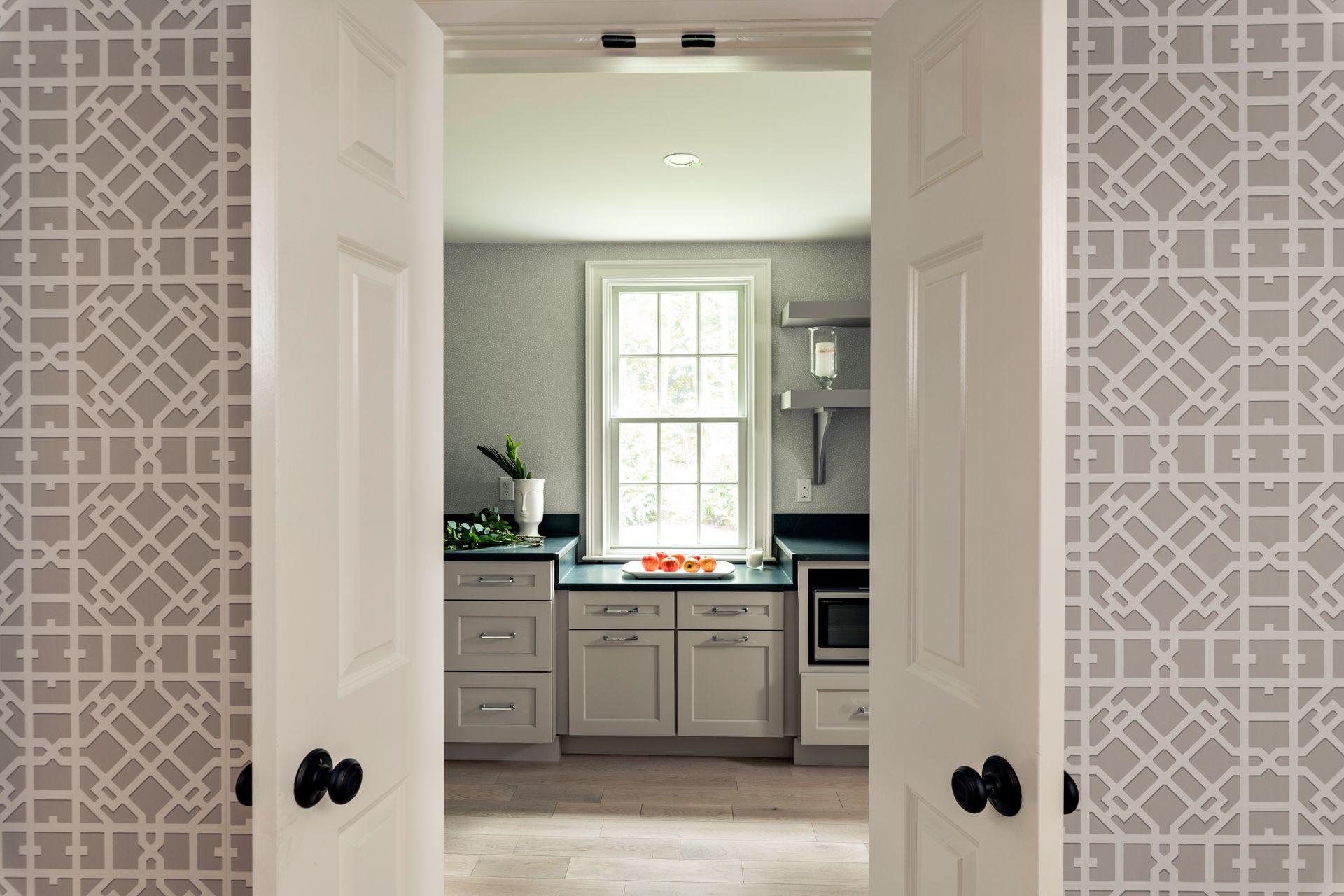 View through open doors into a bright white kitchen with a window over the sink and patterned wallpaper.