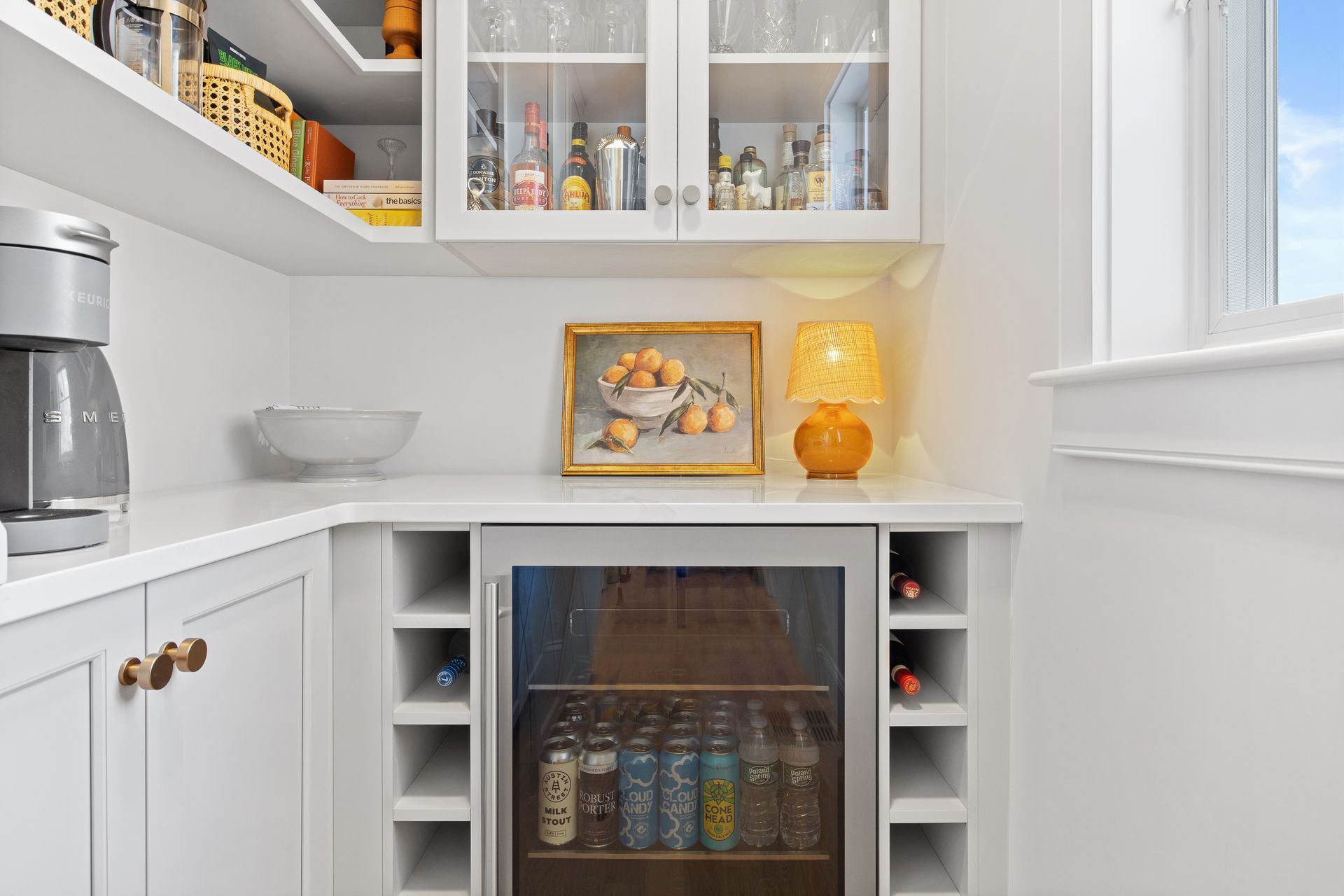 Bright white pantry with cabinets, mini fridge, fruit bowl, lamp, and beverage cooler under a window.