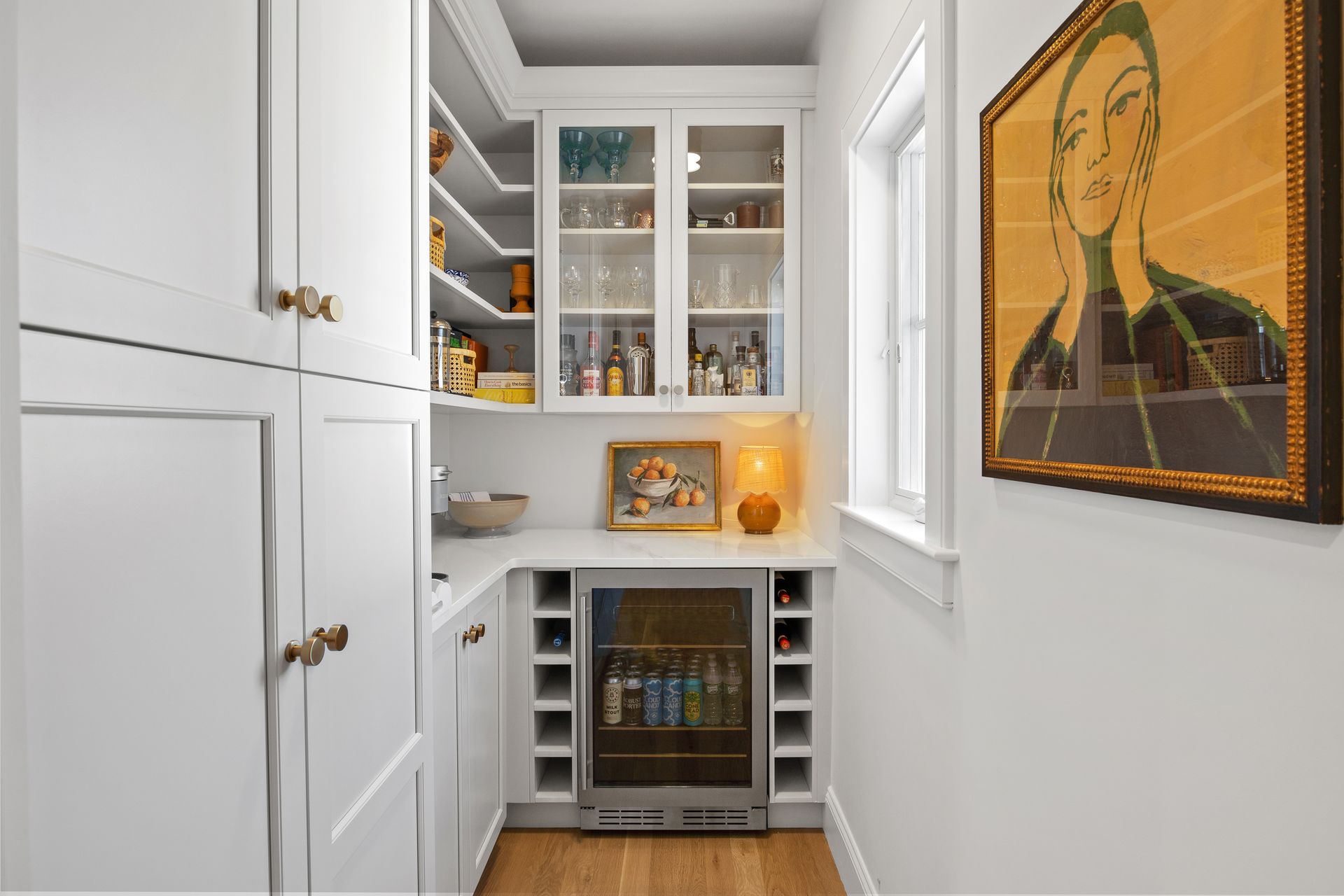 Narrow white pantry with glass cabinets, wine fridge, wood floor, and a framed portrait on the wall.