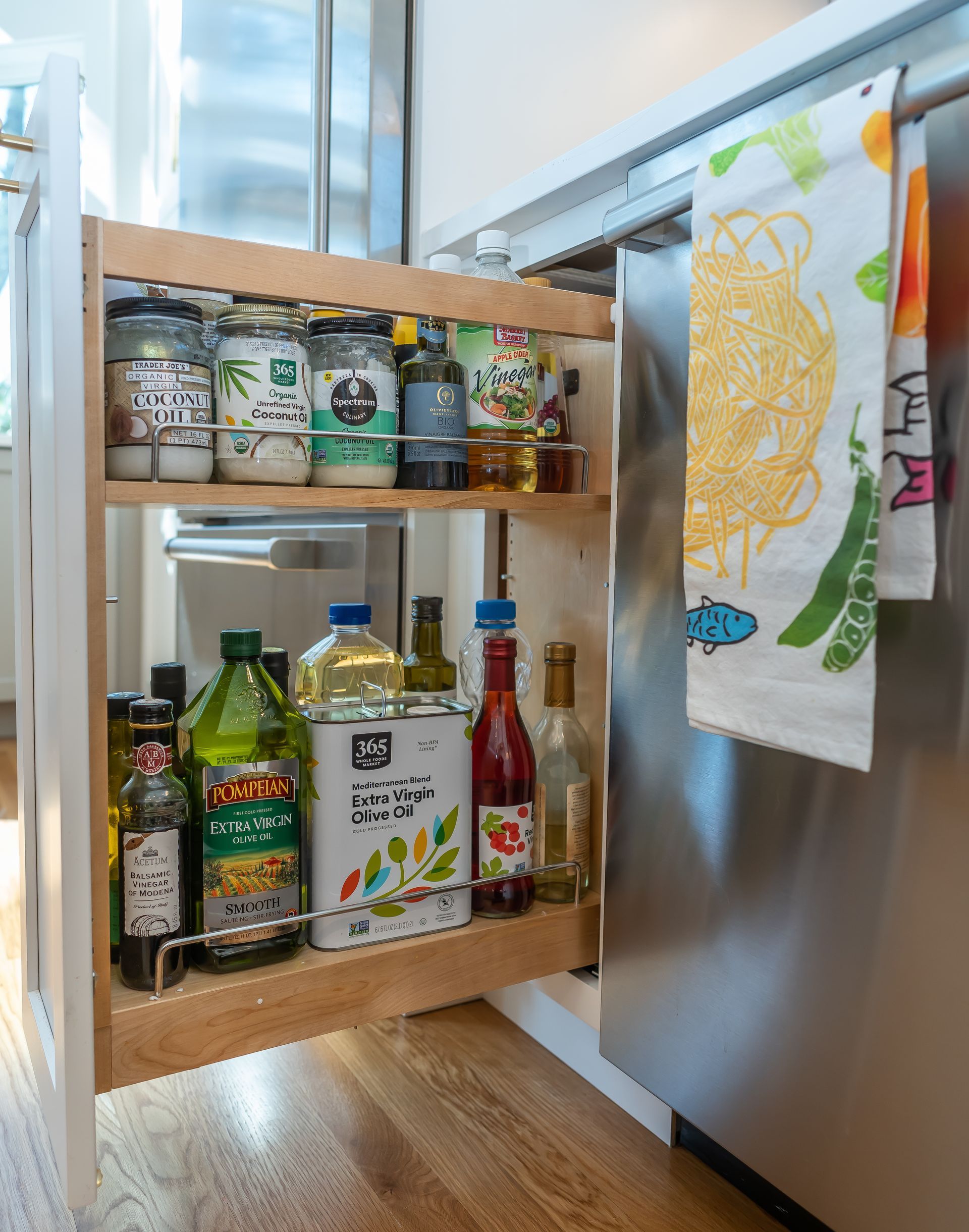 Pull-out pantry shelf with assorted bottles and jars beside a stainless steel refrigerator in a kitchen.