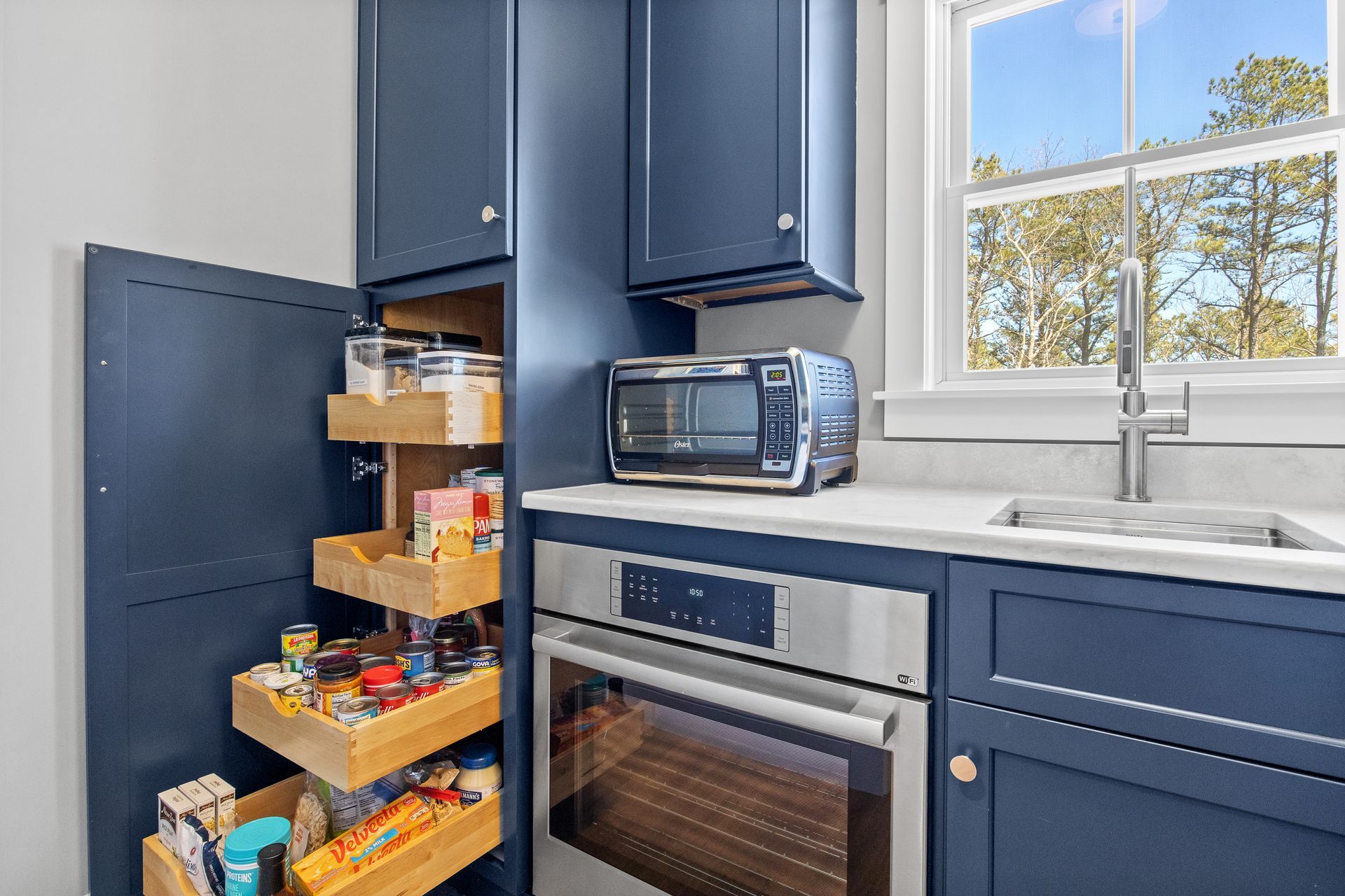 Kitchen with pull-out pantry drawers, dark blue cabinets, microwave, oven, and a window over the sink.