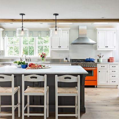 Bright modern kitchen with white cabinets, orange stove, marble island, and fruit bowl by a window