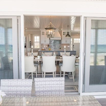 Bright coastal kitchen and dining area viewed through open sliding glass doors with white chairs and bar stools