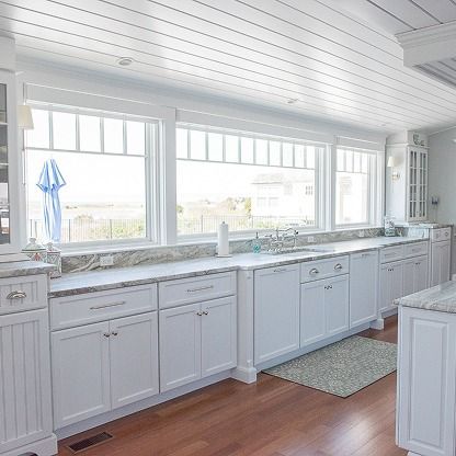 Bright white kitchen with long windows, marble counters, and white cabinets over a wood floor.
