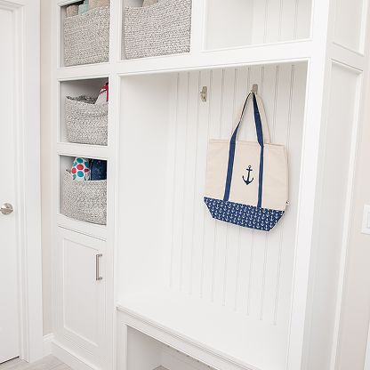White mudroom cubby with woven baskets and a navy anchor tote hanging on a paneled wall