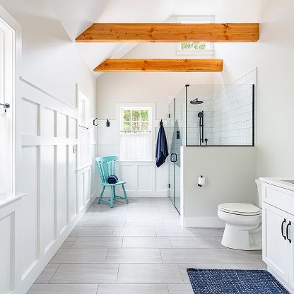 Bright white bathroom with wood ceiling beams, glass shower, toilet, vanity, and a turquoise chair near the window.