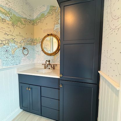 Blue bathroom vanity with white sink, round gold mirror, and dark built-in cabinet beside patterned wallpaper.