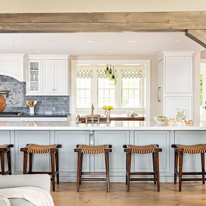 Bright kitchen with white cabinets, blue island, wood beams, and five barstools by a windowed dining nook