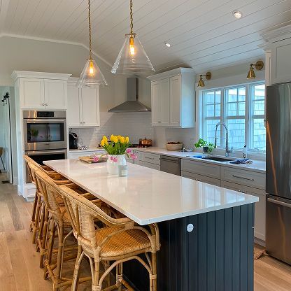 Bright white kitchen with a large island, wicker stools, pendant lights, and a window over the sink.