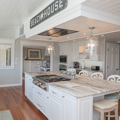Bright coastal kitchen with white cabinets, marble island, pendant lights, and a “BEACH HOUSE” sign